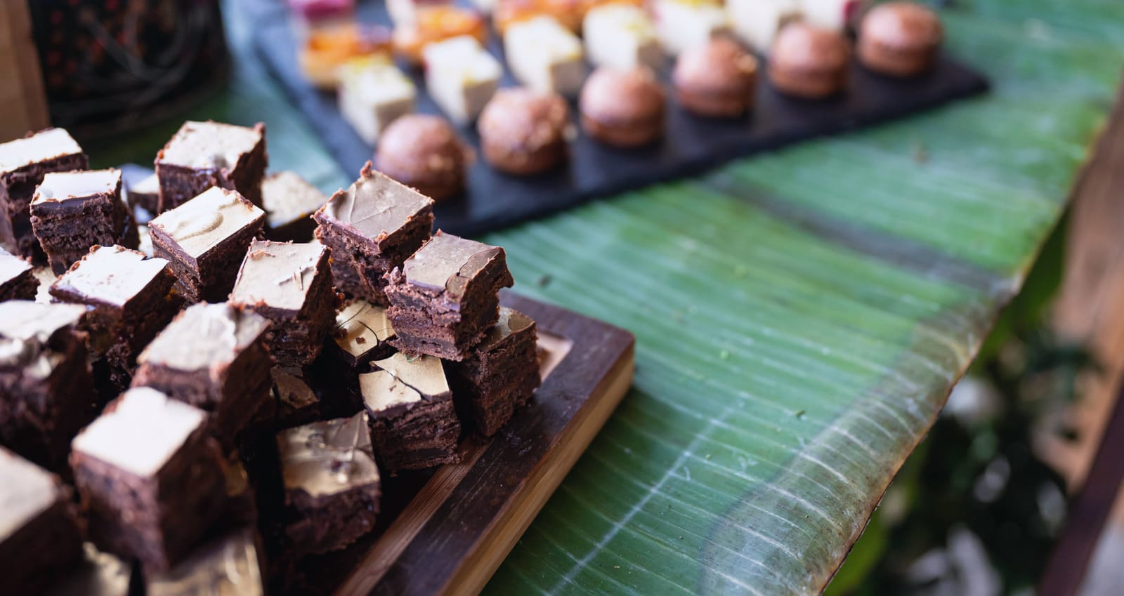 Brownie-Würfel auf einem Holzbrett, dahinter Pralinen und Petits Fours auf Schiefer, ein Bananenblatt als Tischläufer