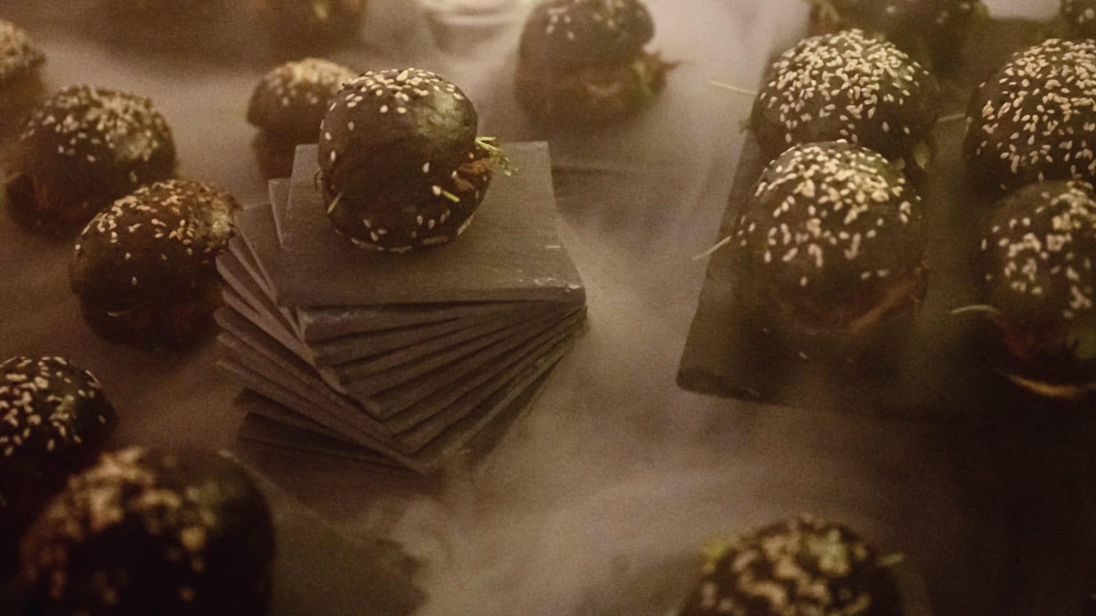 Viele Mini-Burger mit schwarzem Brot und Sesam auf Schieferplatten, dazwischen Nebel