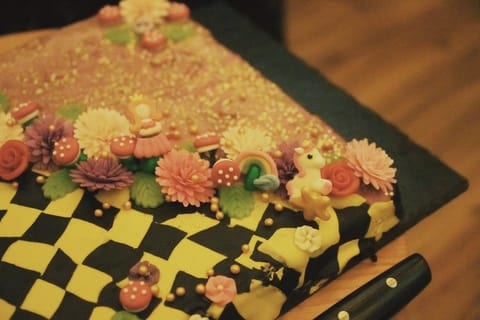 Torte mit schwarz-gelbem Schachbrettmuster, verziert mit bunten Blüten, Pilzen und kleinen Figuren