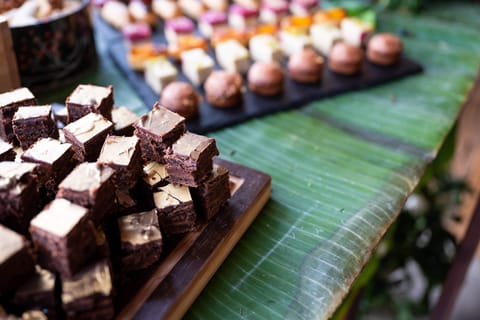 Brownie-Würfel auf einem Holzbrett, dahinter Pralinen und Petits Fours auf Schiefer, ein Bananenblatt als Tischläufer