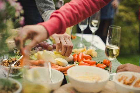 Hände greifen im Freien nach Gemüse auf einer gedeckten Tafel, daneben Schalen und gefüllte Sektgläser