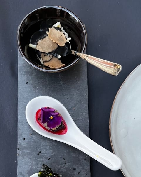 Schwarze Schale mit dunklen Scheiben und einem Löffel, darunter ein Porzellanlöffel mit einer violetten Blüte auf roter Sauce