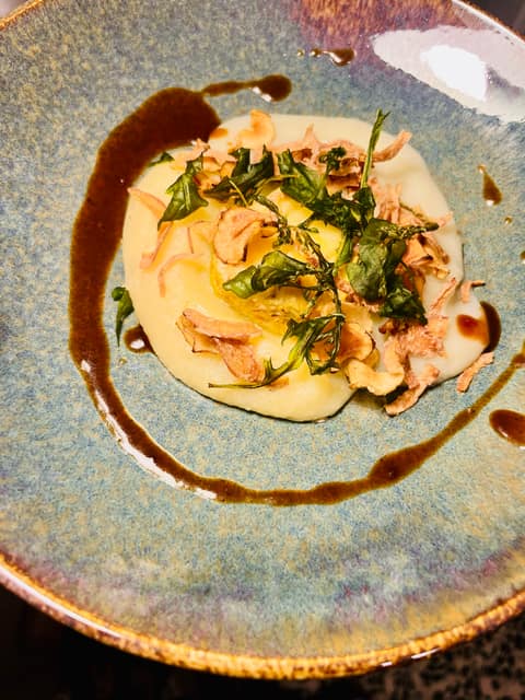 Helles Gericht mit frittierten Kräutern und knusprigen Zwiebeln, umrahmt von einem Schwung dunkler Sauce auf einem blauen Steingutteller