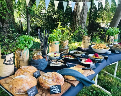 Buffet im Garten unter Wimpeln: Brotlaibe, Kräutertöpfe und Schalen auf blauem Tischtuch