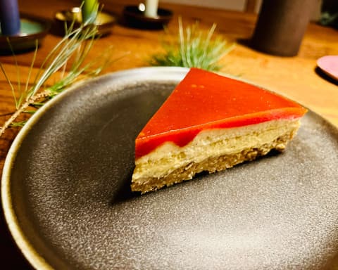 Ein Stück Torte mit roter Glasur auf einem dunklen Teller, daneben Gräser