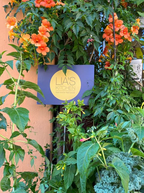 Schild mit dem runden LIA'S KITCHEN-Logo zwischen orangen Blüten und Grün