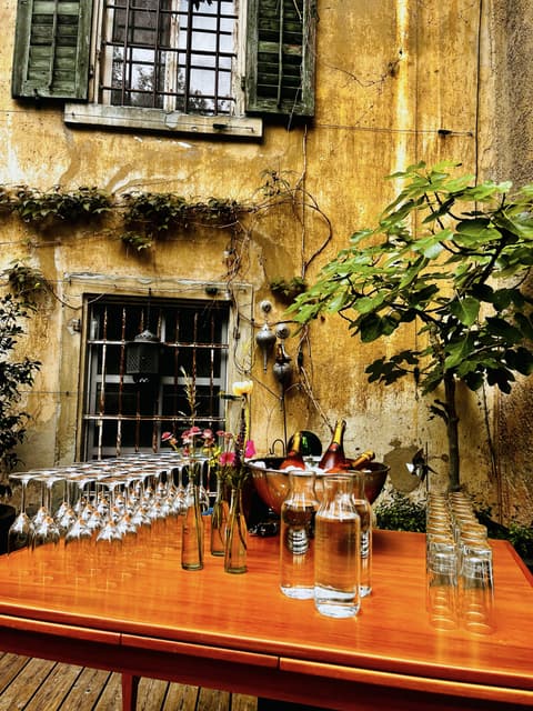 Getränketisch im Innenhof vor einer alten gelben Hauswand: Gläser in Reihen, Wasserkaraffen, Blumen und Wein im Kühler unter einem Feigenbaum