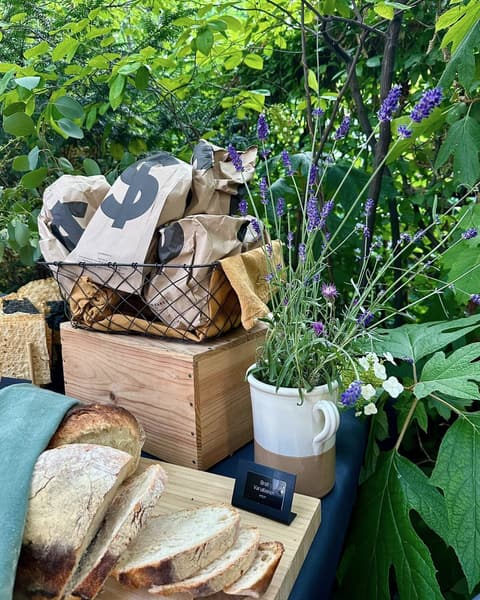 Brotstation im Garten: aufgeschnittenes Brot auf dem Holzbrett, Papiertüten im Drahtkorb und ein Krug mit Lavendel