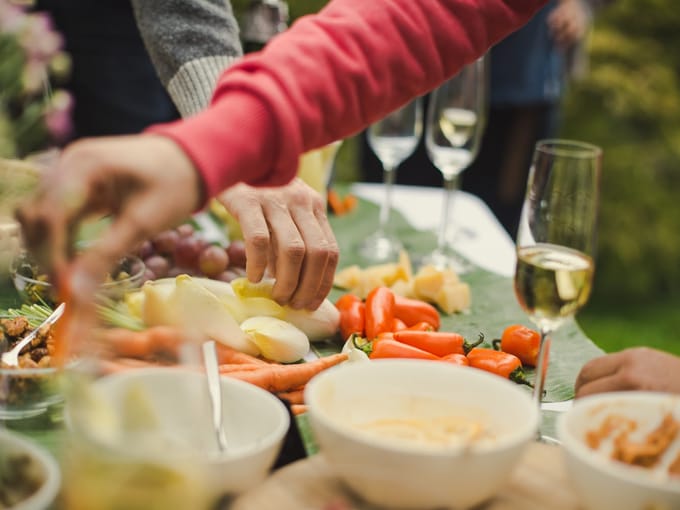 Hände greifen im Freien nach Gemüse auf einer gedeckten Tafel, daneben Schalen und gefüllte Sektgläser