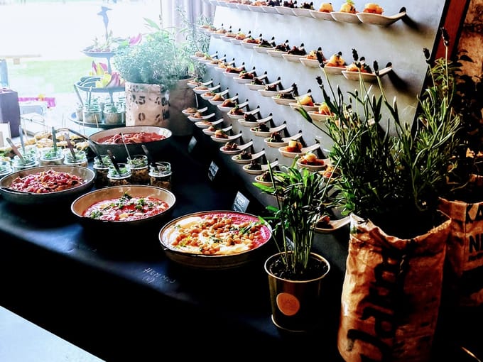 Langes Buffet vor einer Wand voller Porzellanlöffel mit Häppchen, davor Schalen mit Salaten und Kräutertöpfe