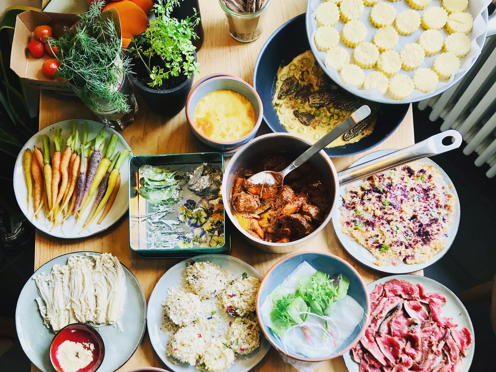 Ein Tisch von oben voller frischer Zutaten und Speisen: bunte Karotten, Kräuter, Tomaten, Schalen und Pfannen